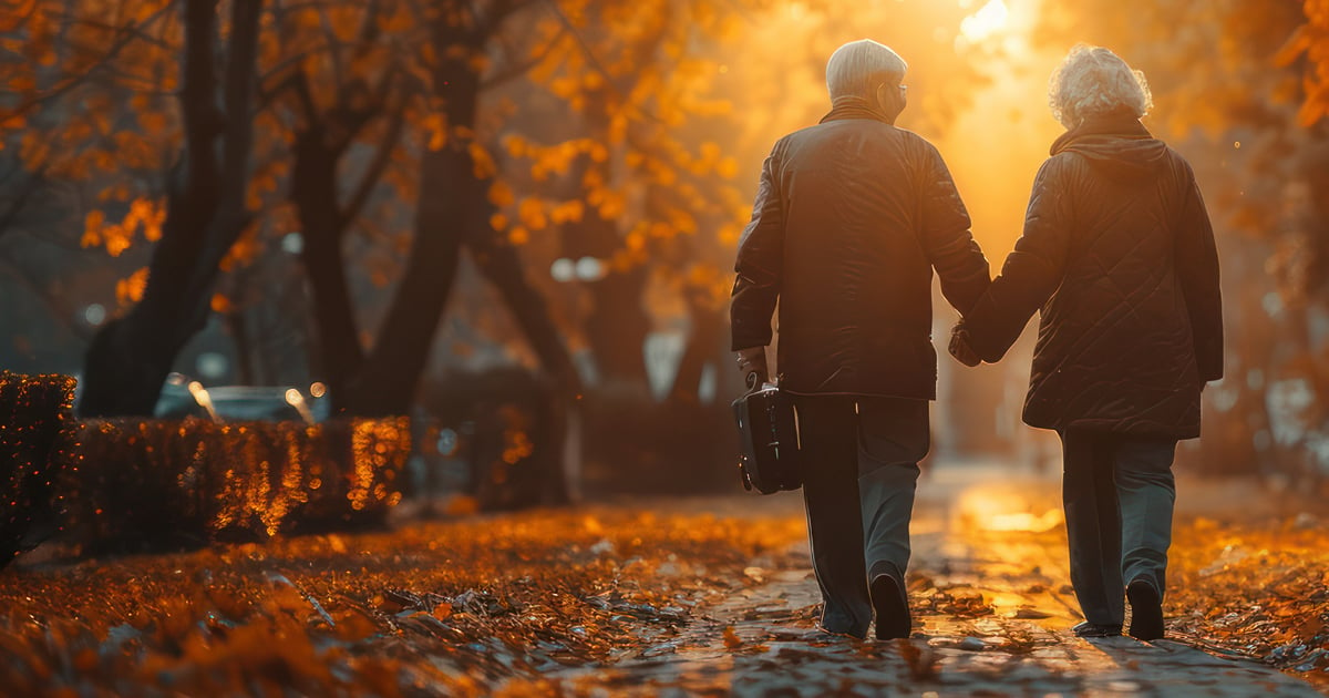 An older couple holding hands and walking down a tree-lined street in autumn.