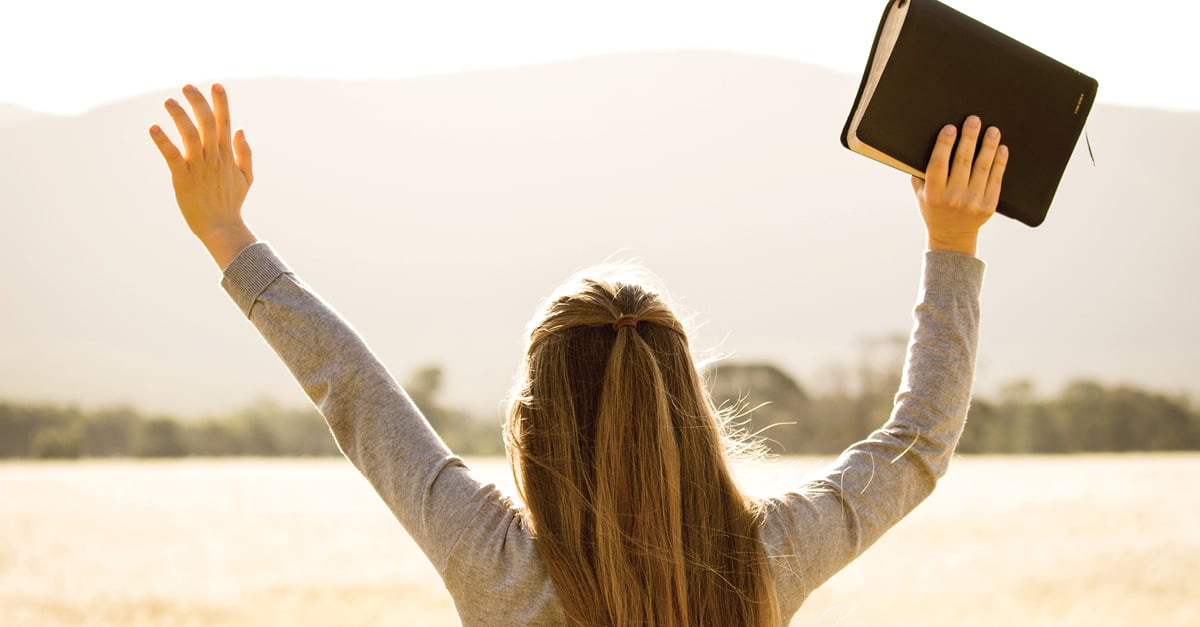 Daily devotional | A woman at a lake, holding a Bible with her arms up.